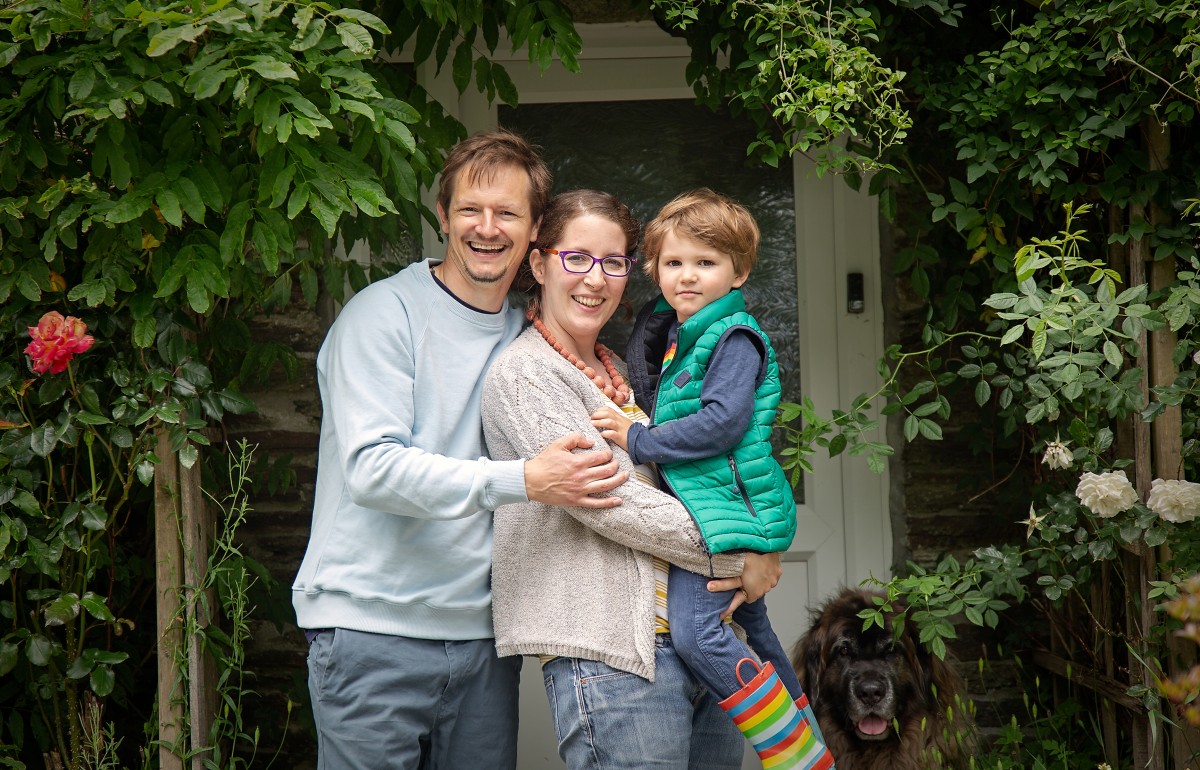 Family photo showing a white man, woman and three year old boy in front of their door surrounded by greenery.