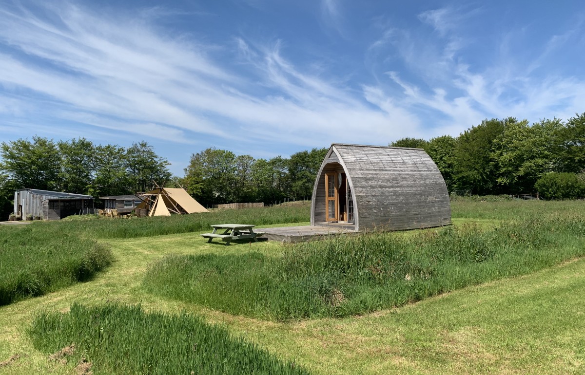 The best camping pod in Cornwall, in front of a blue sky with fire pit area in back