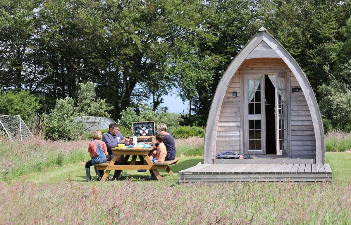 A family sit around a picnic table next to a glamping pod on a site in Cornwall