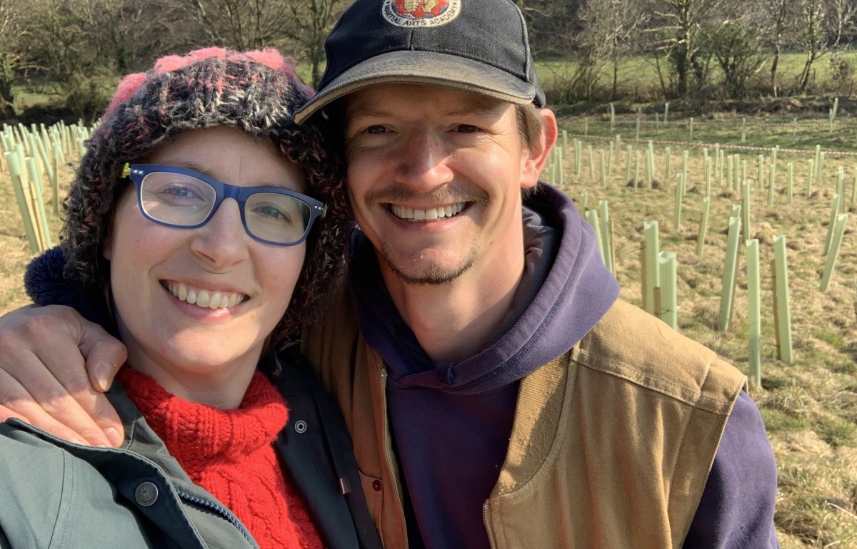 Spencer and India, a white couple in their 40s smile at the camera in their newly planted coppice field.